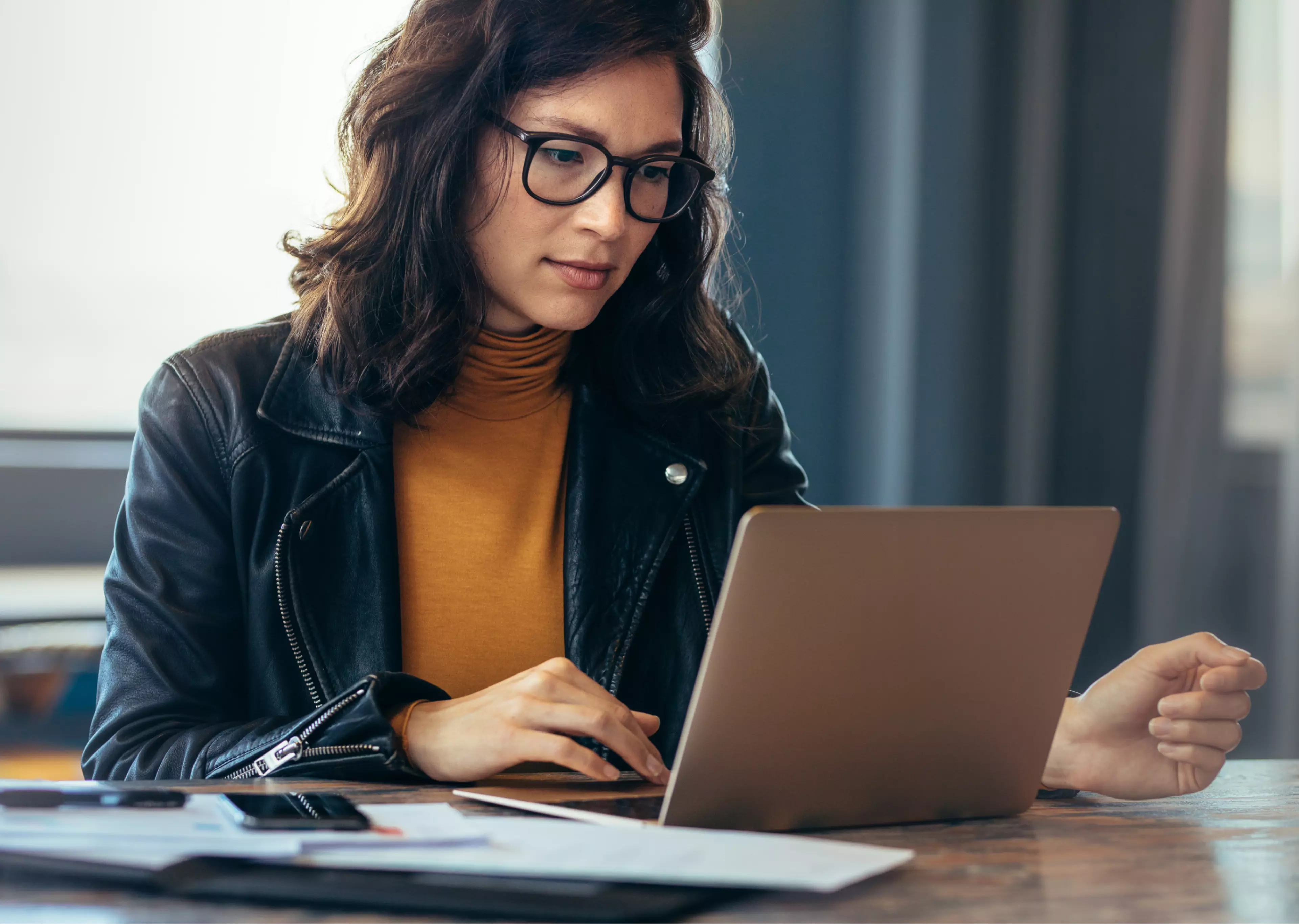 Dark hair woman wearing glasses, sitting behind a desk and looking at her laptop.