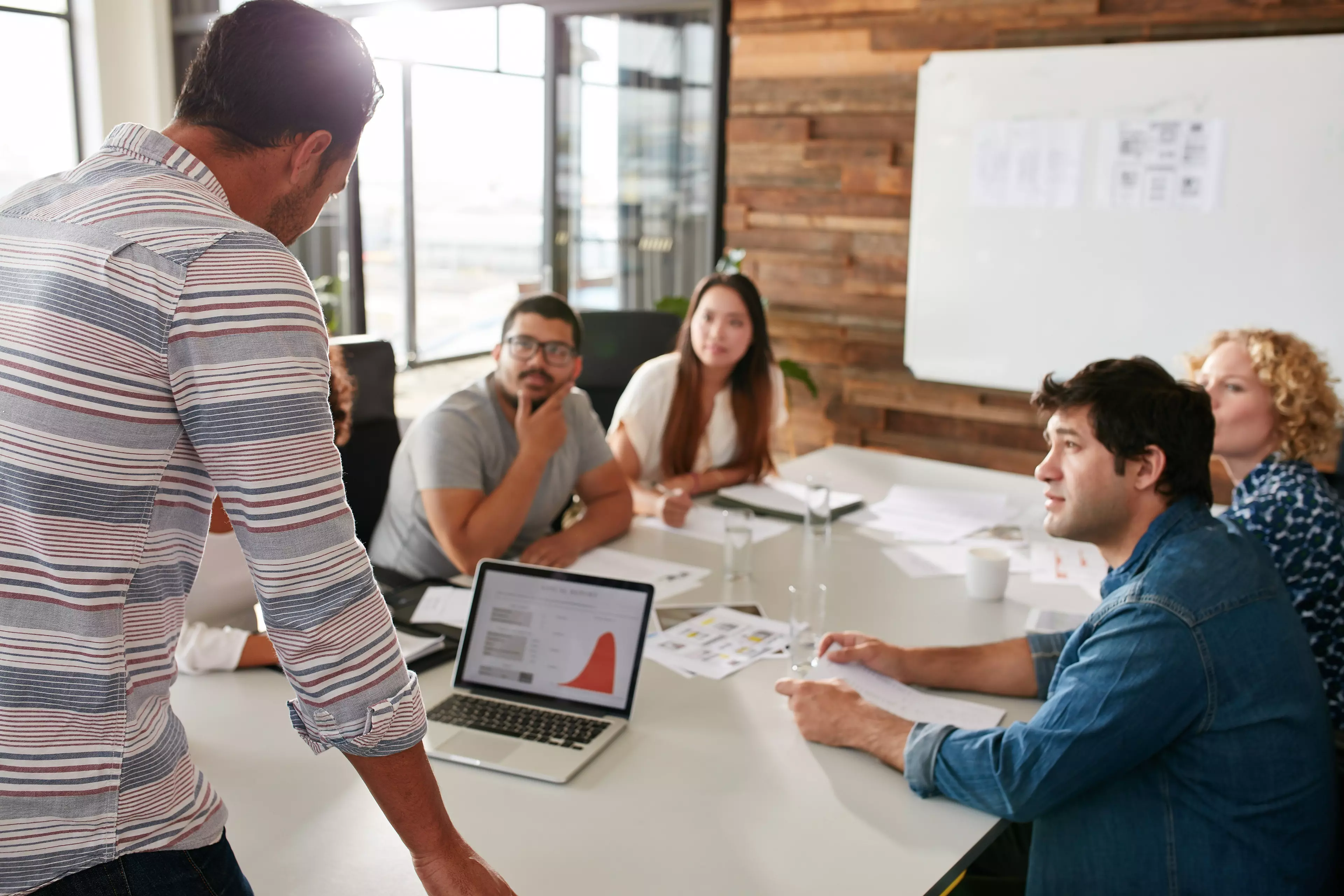 A meeting in the office with 5 people sitting at a table and one man standing and talking to them.