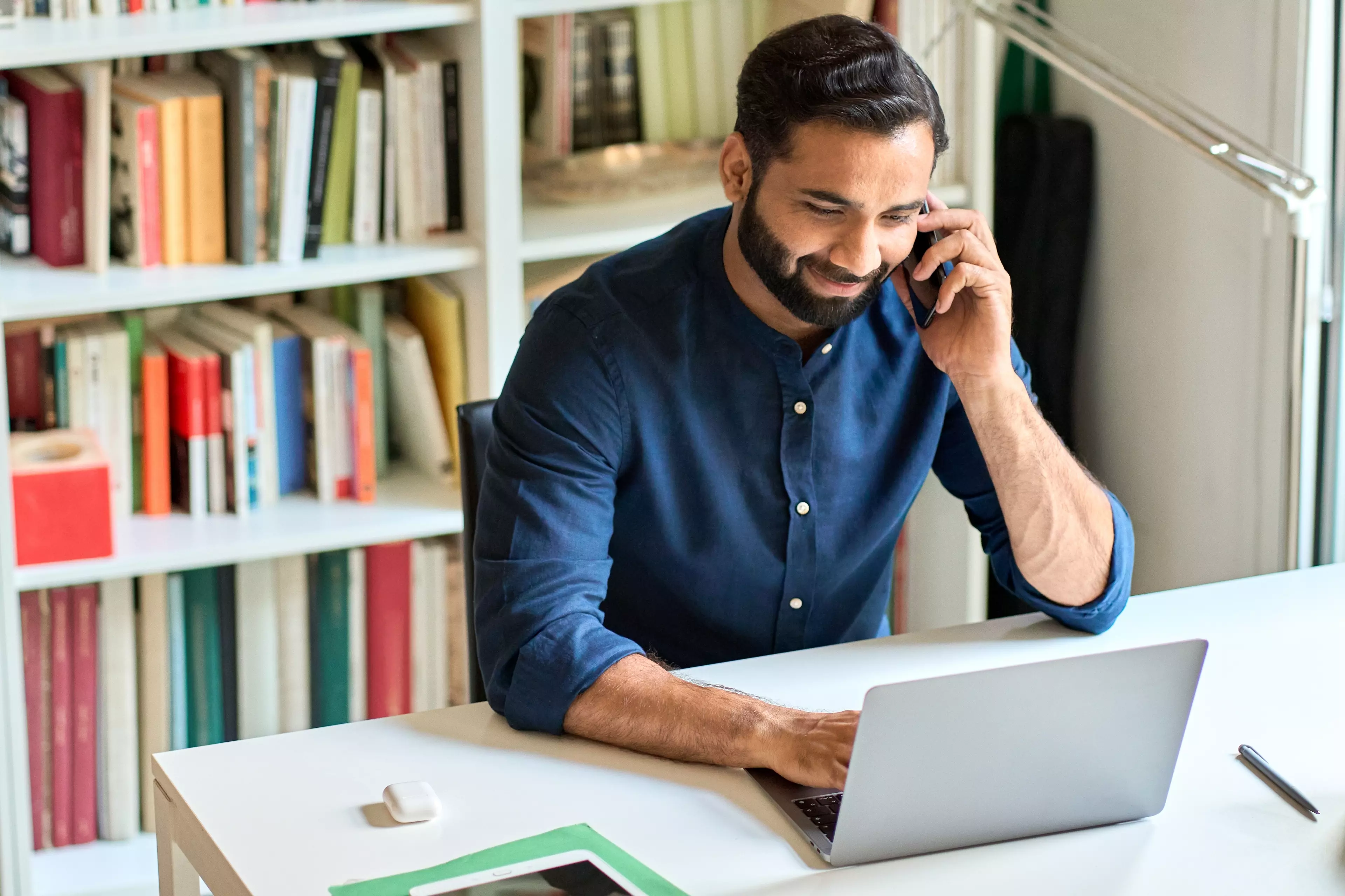 Dark-haired, smiling man sitting behind a desk, working on his laptop and talking over a phone.