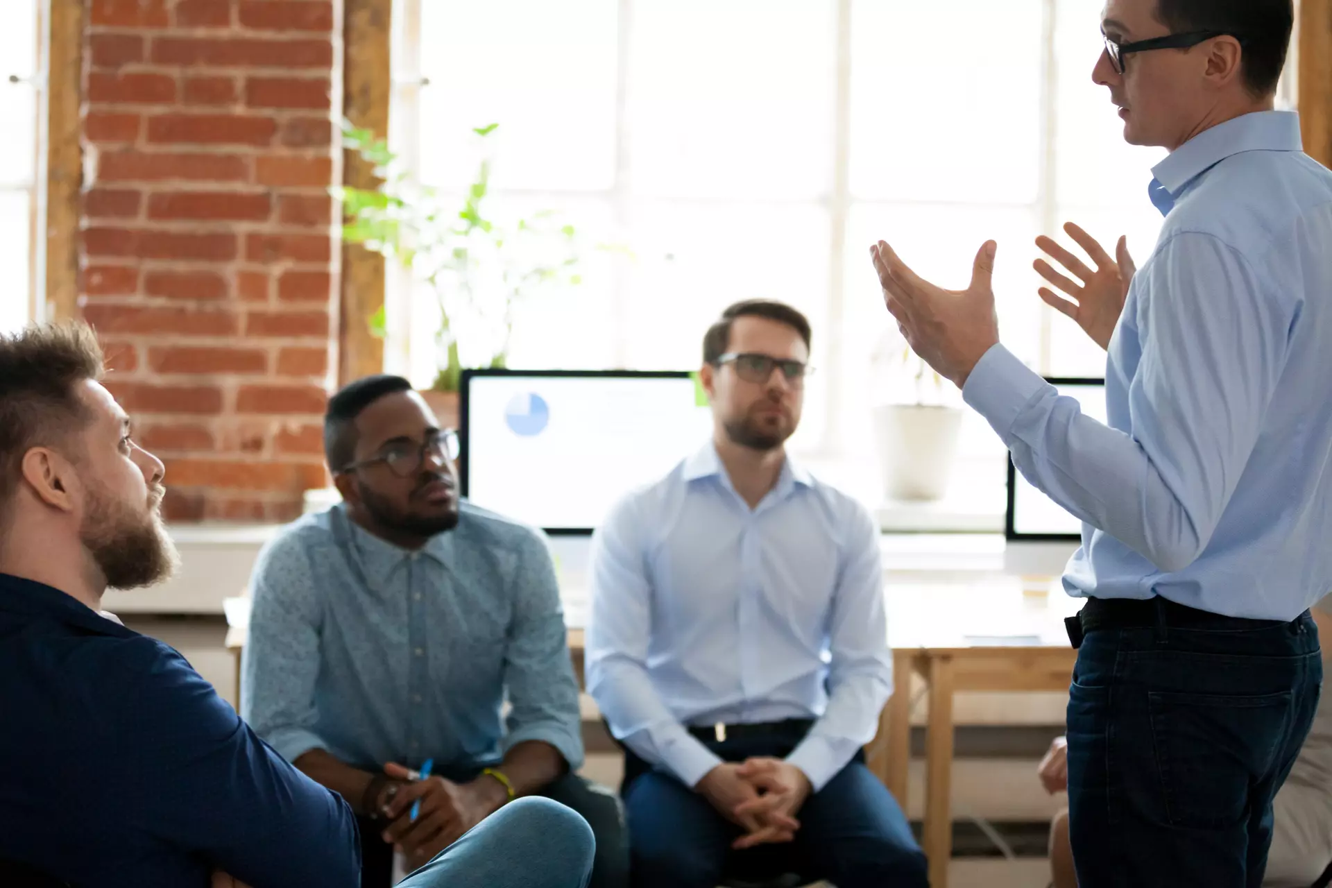 A standing man talks to his sitting colleagues in an office.