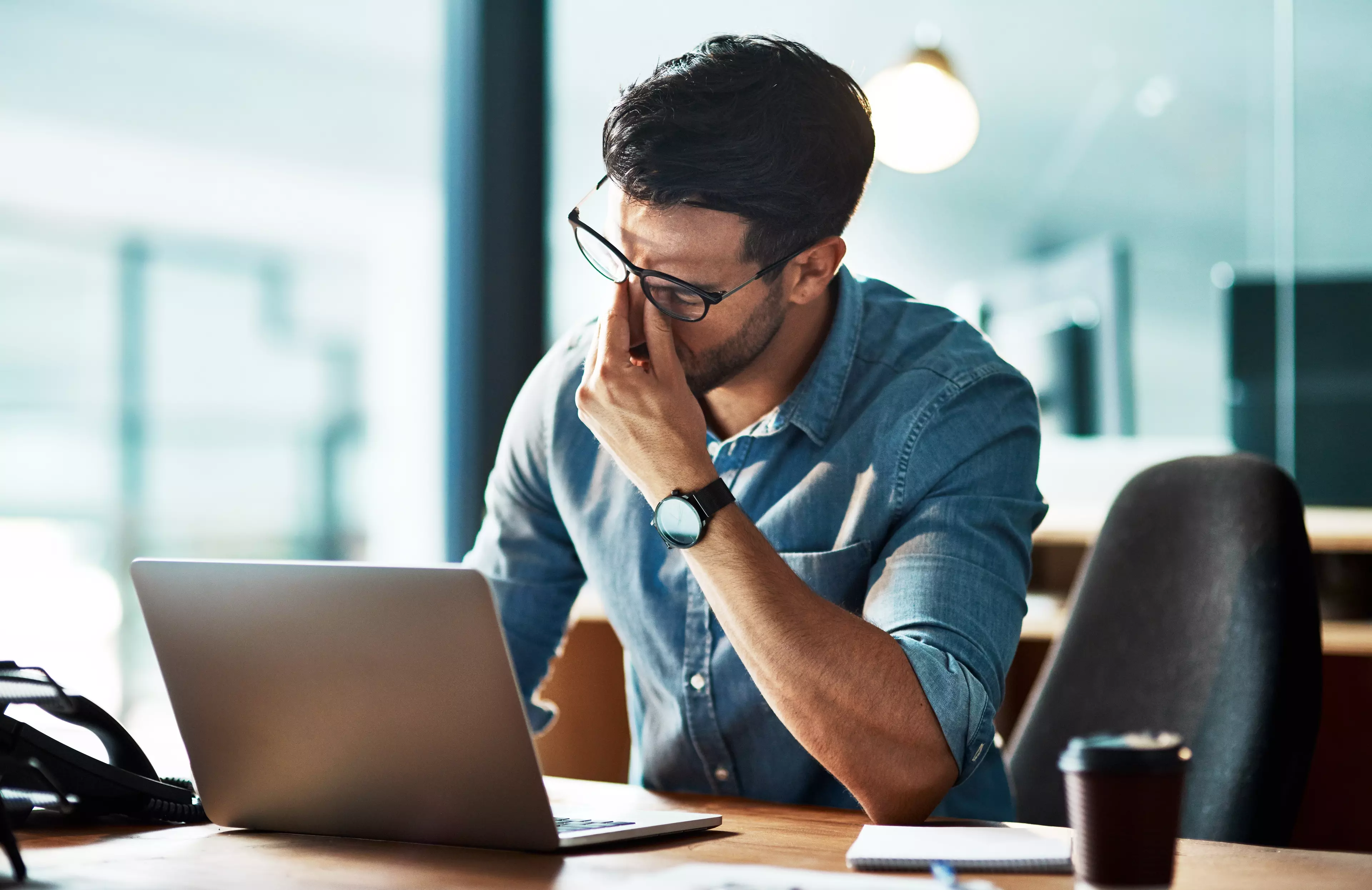 A tired and frustrated man sitting behind a desk and in front of an open laptop.