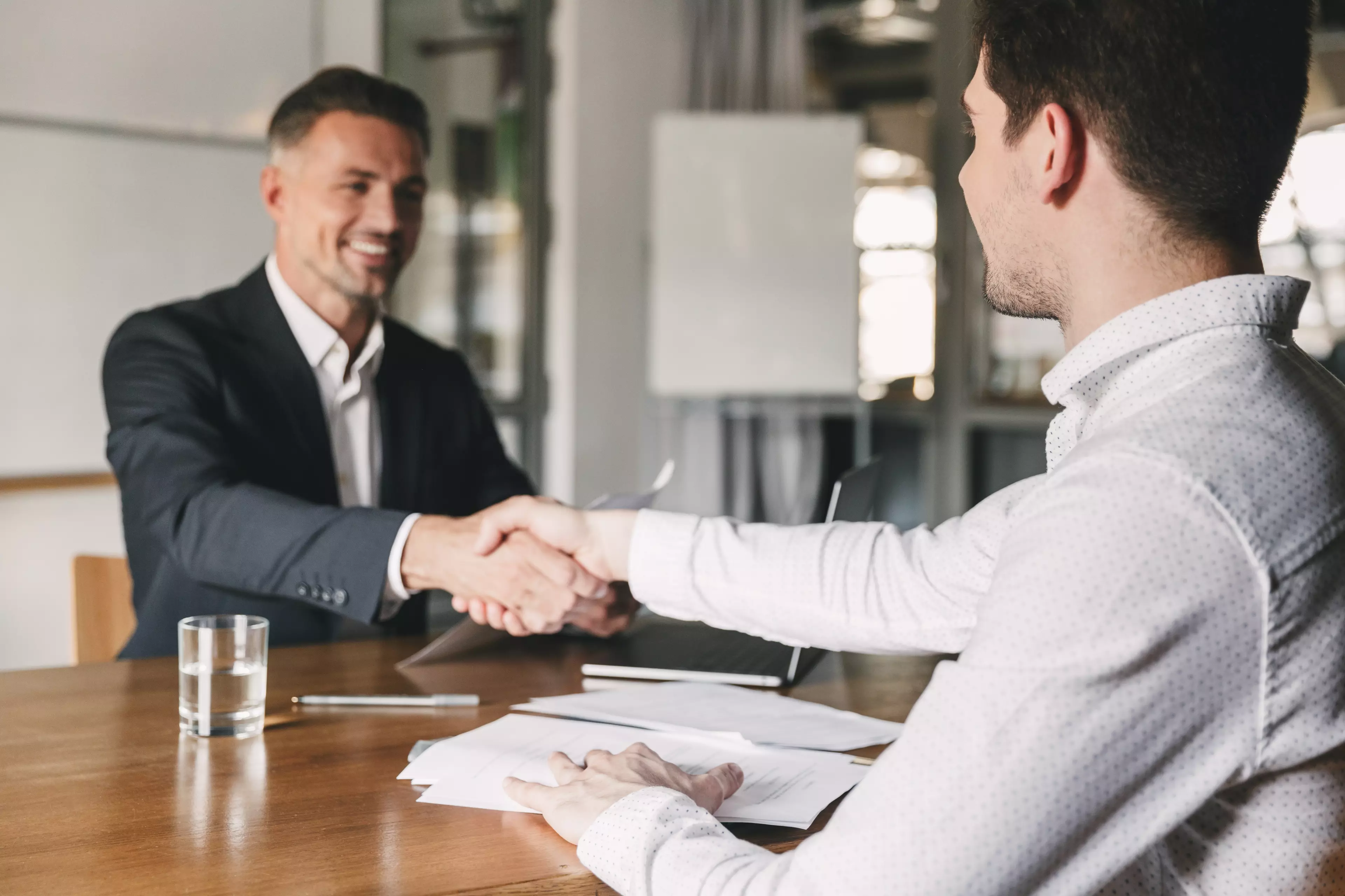 A recruiter and a job candidate sitting  facing each other at a desk, shaking hands.