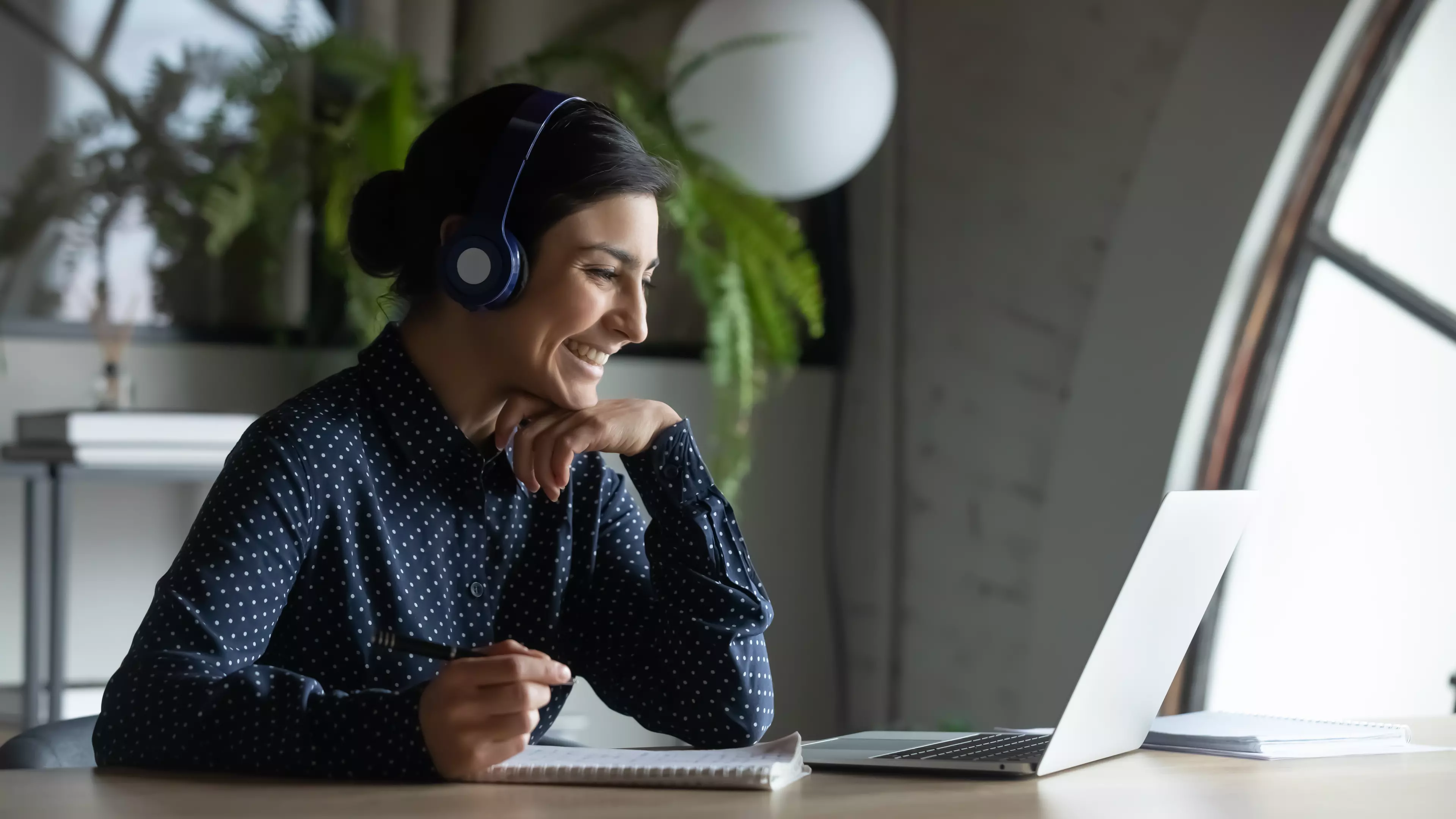 Young, smiling woman sitting behind a desk, talking over videoconferencing software on her laptop.