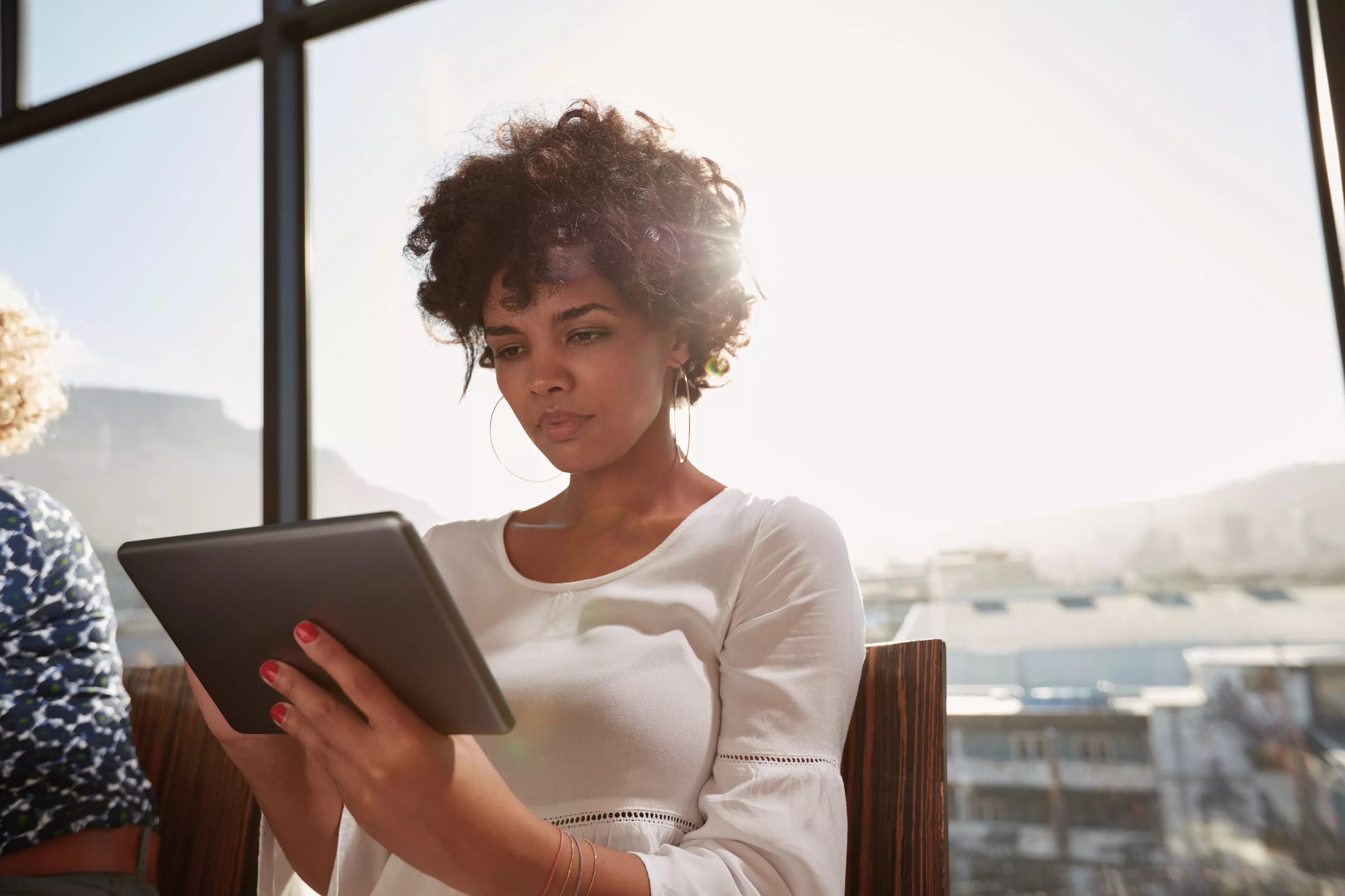  Young, dark-haired woman sitting by a large window, looking at a tablet.