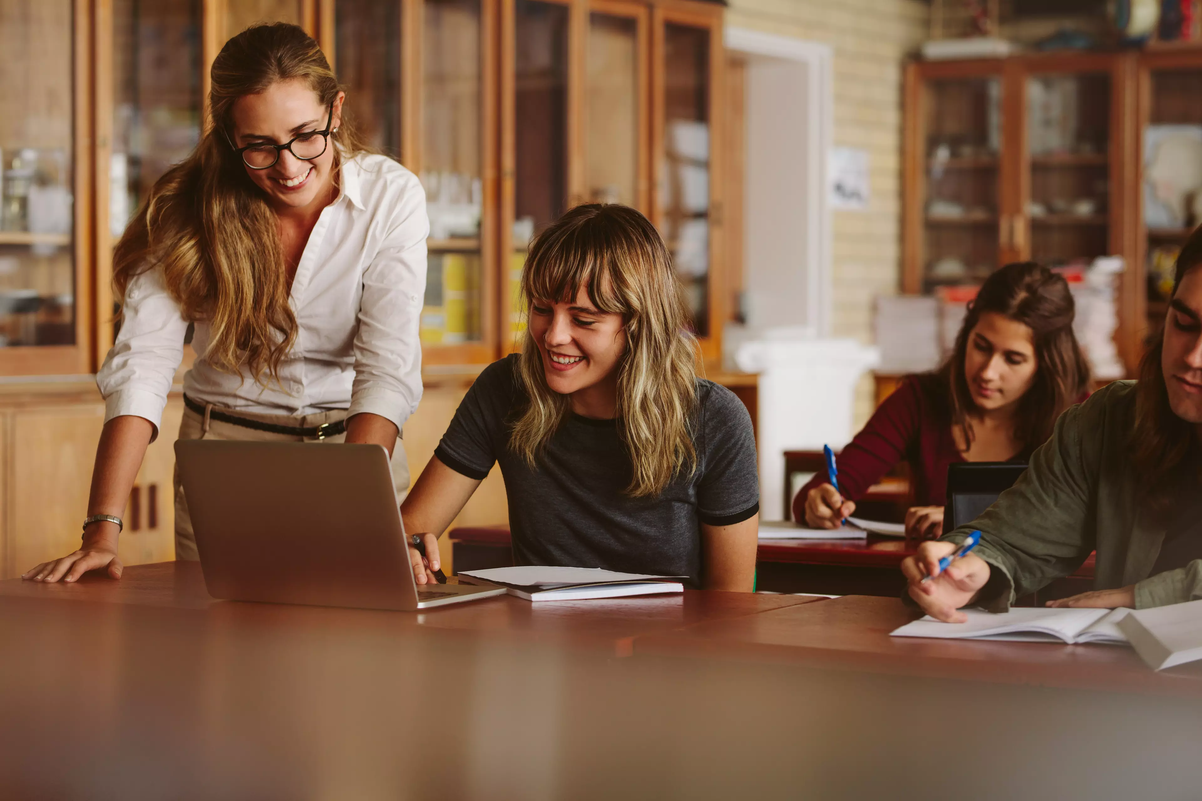 A smiling teacher looks at student's laptop in a classroom.