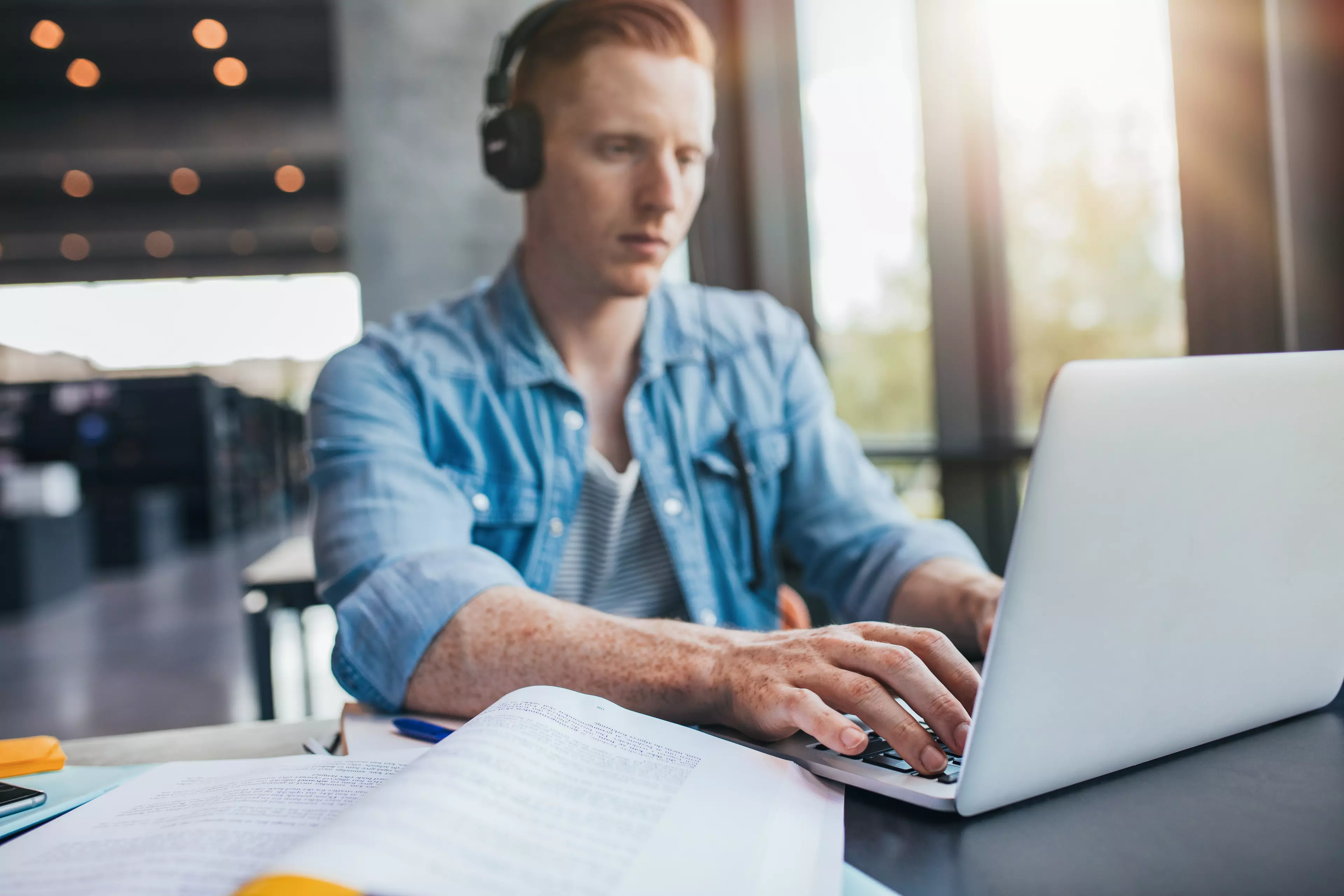  Young, readhead man sitting in front of a laptop, with his headphones on.